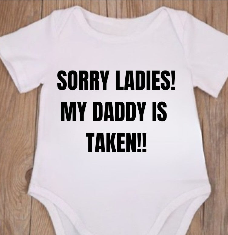 Funny baby onesie with the text 'SORRY LADIES! MY DADDY IS TAKEN!!' on a wooden background.