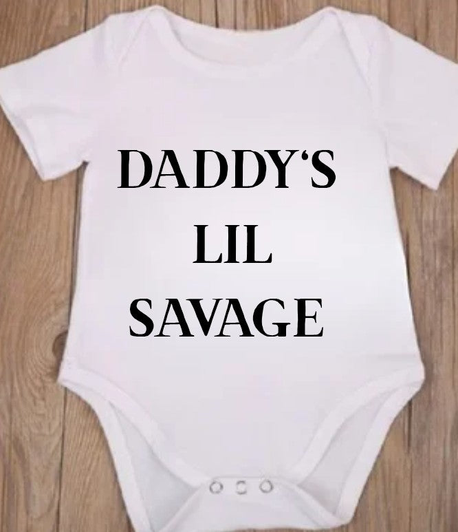 Funny baby onesie with the text 'Daddy's Lil Savage' on a wooden background