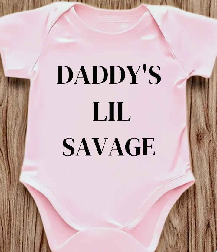 Funny baby onesie with the text 'Daddy's Lil Savage' on a wooden background