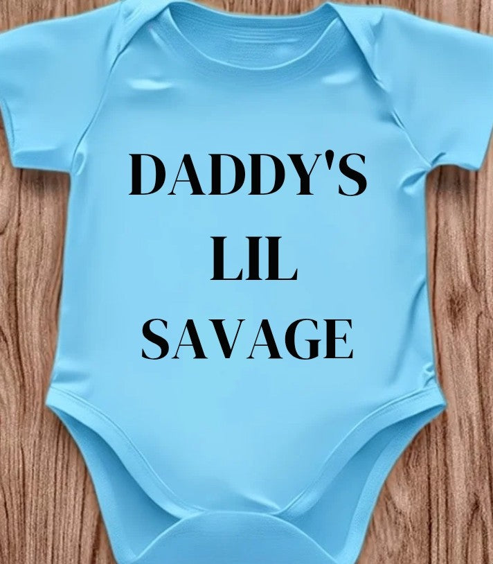 Funny baby onesie with the text 'Daddy's Lil Savage' on a wooden background